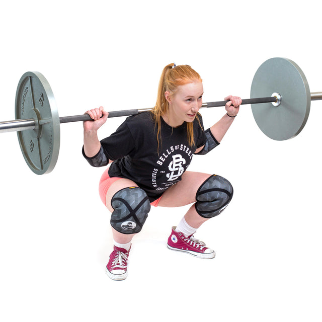 A woman with long red hair does a barbell squat wearing a black shirt, pink shorts, Bells of Steel Knee Sleeves for support, and red sneakers. The background is white and uncluttered.