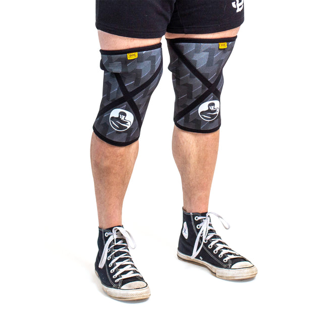 A person wears Bells of Steel Knee Sleeves featuring a white graphic, paired with black-and-white high-top sneakers. Only their legs, knees, and feet are shown against a white background.