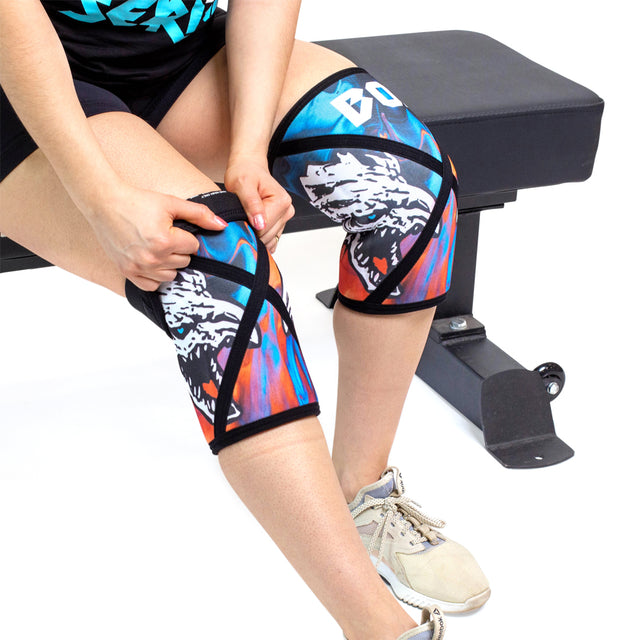 A person on a workout bench adjusts Bells of Steel Knee Sleeves featuring a wolf’s head graphic. Wearing shorts, a black shirt, and light athletic shoes, they prep for serious squat support with these colorful compression sleeves.