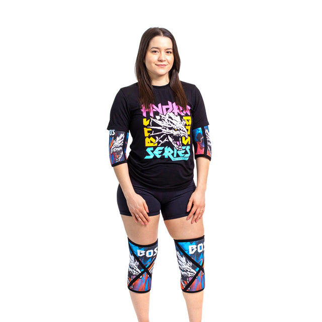 A woman faces forward, smiling against a white background, wearing Bells of Steel Knee Sleeves with bold patterns and "BOSS" on the knees for squat support, along with a black graphic T-shirt and black shorts.