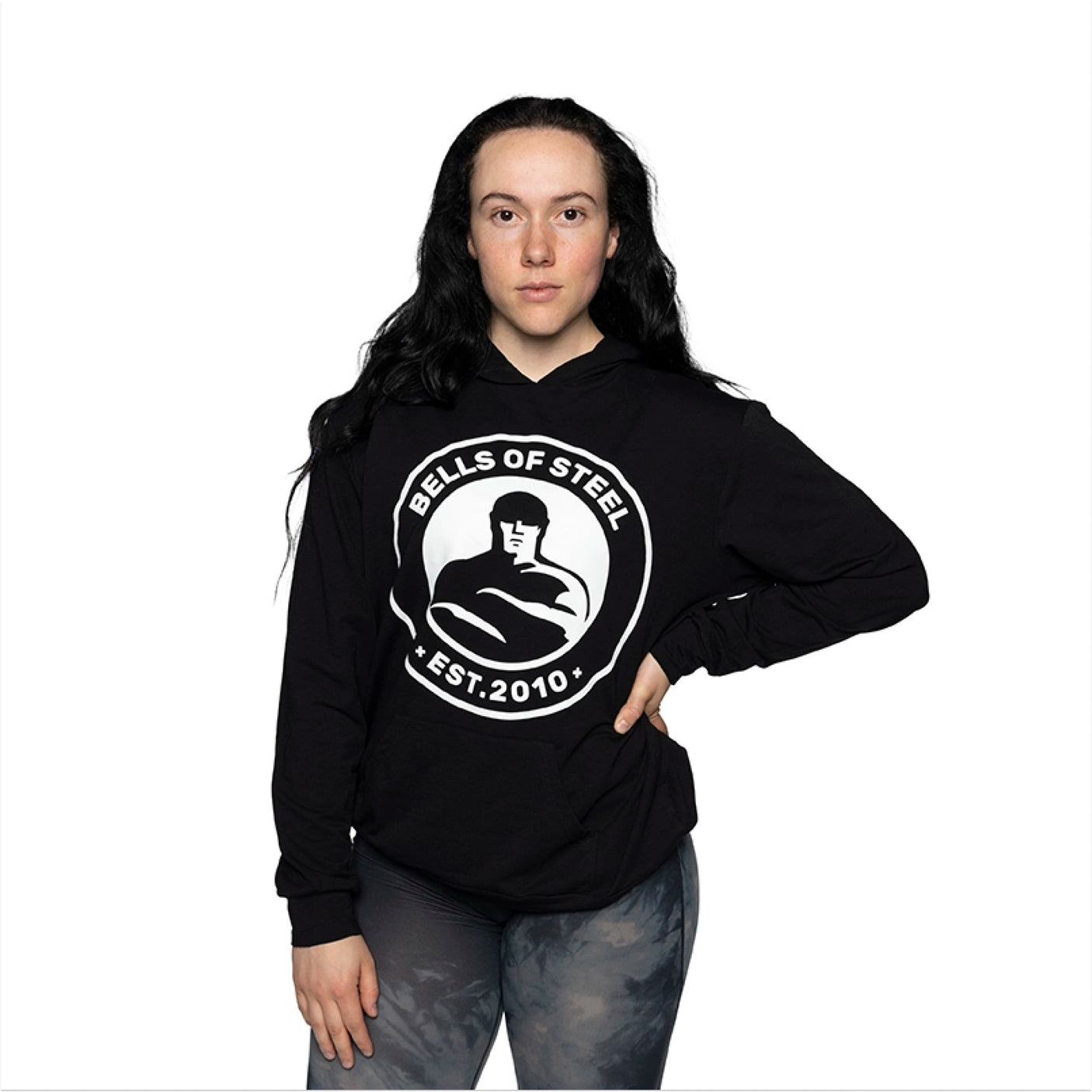 A woman with long dark hair wears the Bells of Steel Bamboo Hoodie, featuring "Bells of Steel Est. 2010" on the front, paired with gray patterned leggings. She stands against a white background with one hand on her hip.