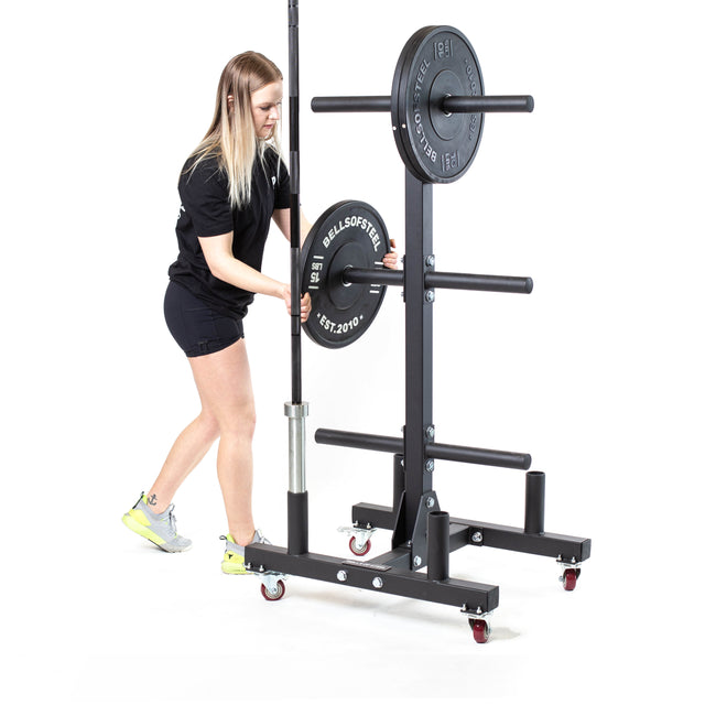 A woman in athletic apparel loads a weight plate onto a barbell held by the Bells of Steel Bumper Plate Weight Tree And Bar Holder, a black wheeled rack with integrated storage, set against a white background.