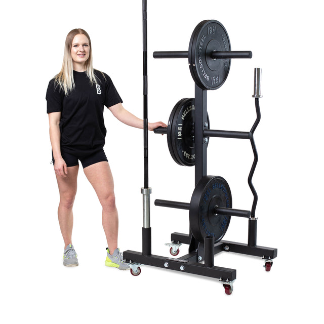 A woman in a black t-shirt and shorts stands beside the Bells of Steel Bumper Plate Weight Tree And Bar Holder, an organized storage rack for barbells and plates. This mobile unit stores standard and specialty bars against a white background.