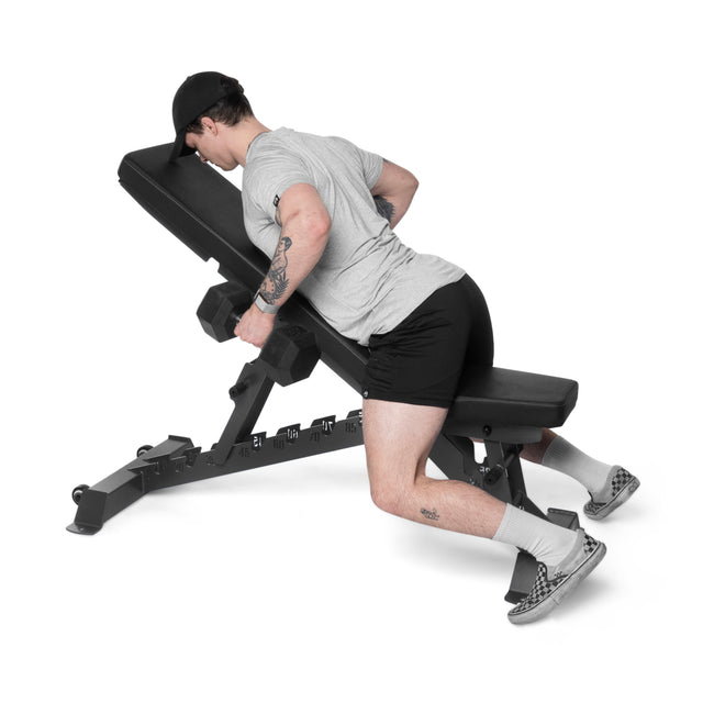A person in gym attire, including a cap, shorts, socks, and shoes, uses the Buzz-Saw Heavy-Duty Adjustable Bench by Bells of Steel. Lying face down with dumbbells in each hand and arms extended forward demonstrates exercise versatility.