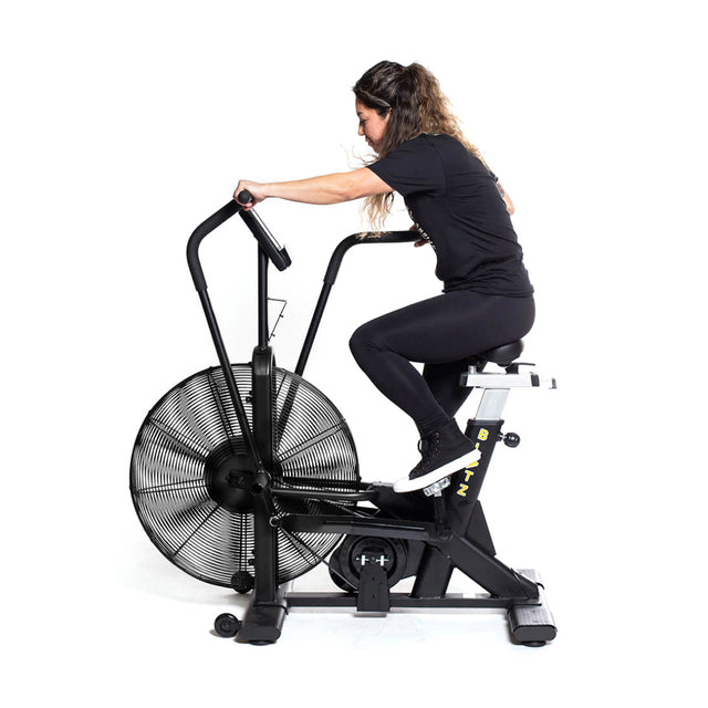 A woman in black athletic wear uses the Bells of Steel Blitz Air Bike, gripping the handlebars and pedaling to demonstrate a full-body workout against a plain white background.