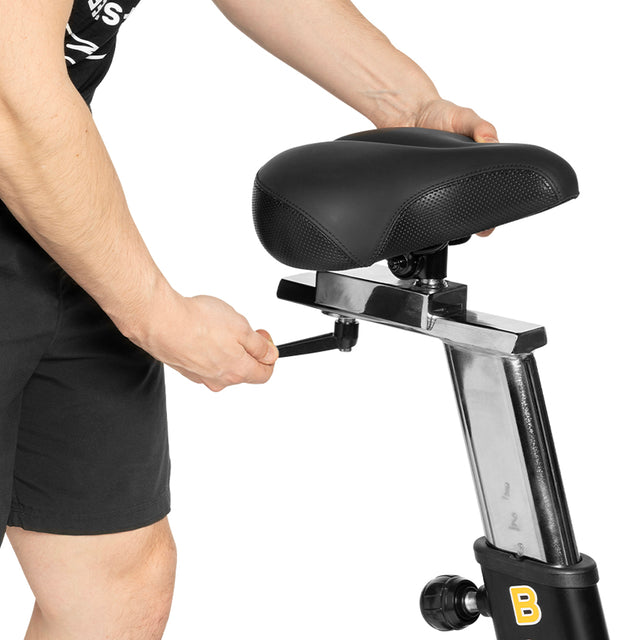A person adjusts the height of the Blitz Air Bike seat by turning a knob on the metal post, getting ready for a full-body workout with the Bells of Steel bike in their home gym.