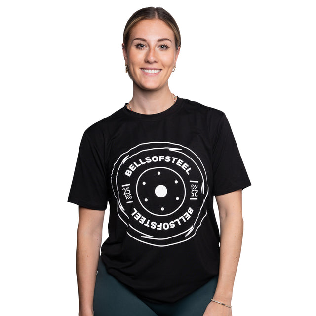 A smiling woman stands against a white background wearing the Bells of Steel Bamboo T-Shirt, a black, moisture-wicking gym shirt featuring a large white weight plate graphic and "BELLS OF STEEL 25 KG" printed on the front.