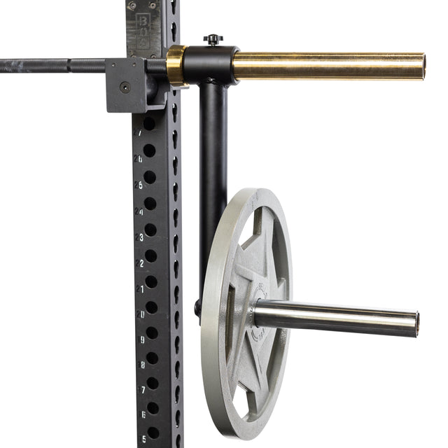 A close-up of the Bells of Steel Camber Bar Attachment - Pair mounted on a gym rack, supporting a large weight plate and an Olympic barbell. The rack features numbered holes for adjustable height.