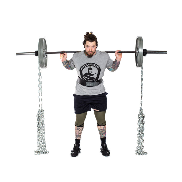 A bearded person with tattoos stands on a white background, wearing a gray shirt, black shorts, knee sleeves, and lifting shoes, holding a barbell loaded with weight plates and Bells of Steel Weightlifting Chains on their shoulders.