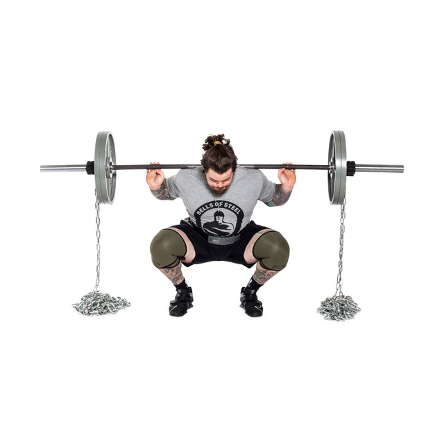 A tattooed person with a bun squats with a barbell loaded with weight plates and Bells of Steel Weightlifting Chain Sets, wearing a gray shirt, black shorts, knee sleeves, and lifting shoes for progressive resistance.