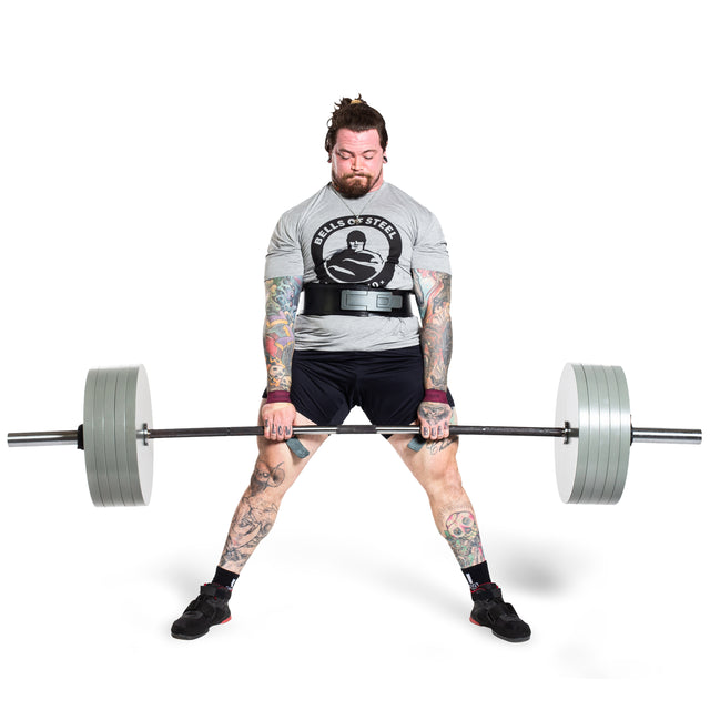A heavily tattooed man in a Bells of Steel shirt and shorts performs a sumo deadlift with a barbell loaded with Bells of Steel Machined Iron Olympic Weight Plates against a white background.