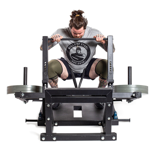 A tattooed man with a man bun grips the handles of a loaded squat machine, fitted with Bells of Steel Machined Iron Olympic Weight Plates. He looks down, wearing a gray T-shirt, shorts, and knee sleeves against a white background.