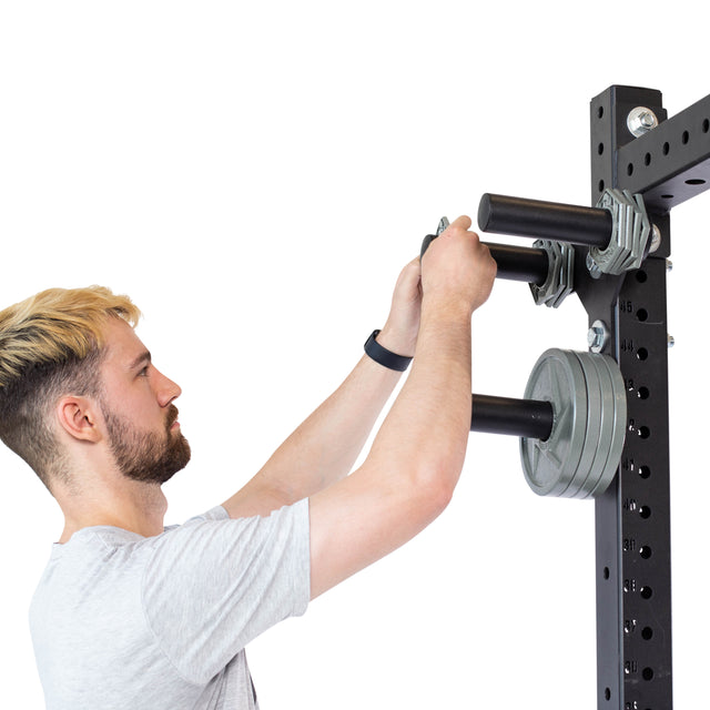 A man with short hair adjusts weight plates on wall-mounted Change Plate Storage Pegs by Bells of Steel. Wearing a light gray T-shirt and a fitness tracker, he arranges Olympic change plates on the black rods, which have multiple holes for precision training.