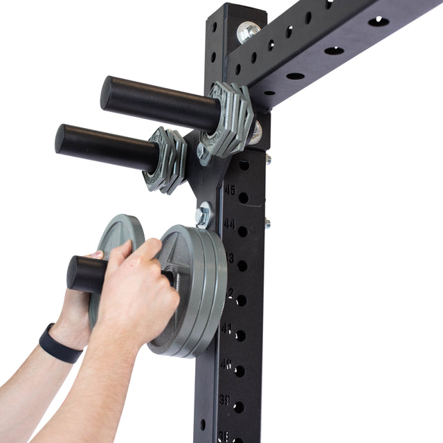 A person methodically arranges Olympic change plates onto Bells of Steel’s black Change Plate Storage Pegs with rods. The rack, featuring multiple holes for versatile organization, stands vertically. Their hands, showing a sleek black wristband, are visible.