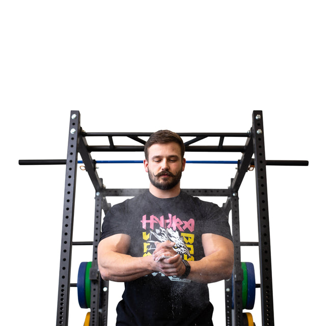 A man stands before a squat rack, clapping his Bells of Steel Mighty Grip Workout Chalk-covered hands as white dust rises. Focused and wearing a black graphic T-shirt, he prepares to lift weights against a plain white background.