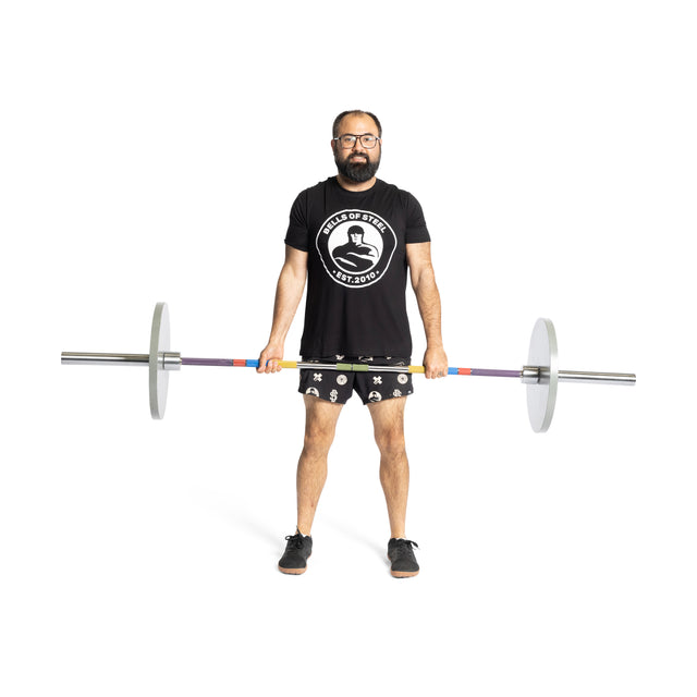 A bearded man in glasses, a black t-shirt, and patterned shorts holds the Bells of Steel Hydra Seven Ring Bar horizontally in front of him against a white background.
