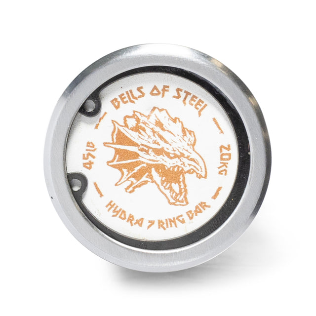A close-up of the Hydra Seven Ring Bar’s weight plate end cap by Bells of Steel, showing an orange dragon head and the text "BELLS OF STEEL HYDRA 7 RING BAR 45LB 20KG" on white with a silver outer ring.