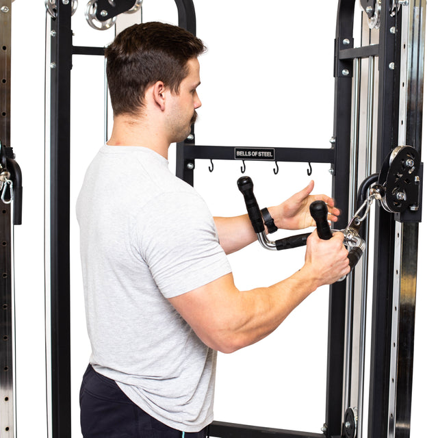 A man in a gray t-shirt stands at a cable machine labeled "Bells of Steel," holding the Multi Purpose Close Grip Cable Attachment with both hands as he prepares to exercise.
