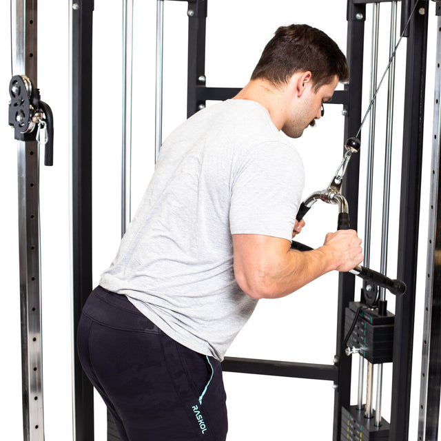 A man in a gray shirt and black pants performs a triceps pushdown on a cable machine, using the Bells of Steel Multi Purpose Close Grip Cable Attachment with both hands while leaning slightly forward.