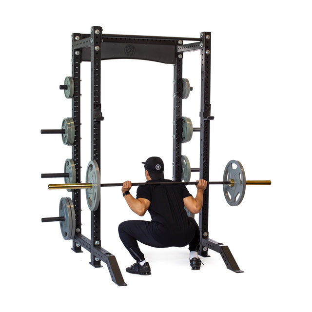 A person in black workout gear and a cap performs a barbell squat inside the Bells of Steel Hydra Collegiate Power Rack (3" x 3", ⅝" holes)—a top choice to inspire your home gym setup.