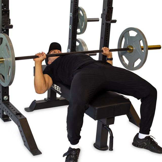 A person in black athletic wear and a hat prepares to bench press with a barbell on the Bells of Steel Hydra Collegiate Power Rack - Prebuilt (3" x 3", ⅝" Holes) in a gym.