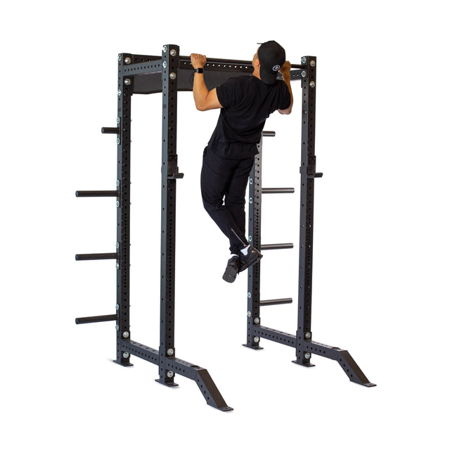 A person dressed in black athletic clothing performs a pull-up on the Bells of Steel Hydra Collegiate Power Rack - Prebuilt (3" x 3", ⅝" Holes) with weight plate storage, perfect for any home gym, against a white background.