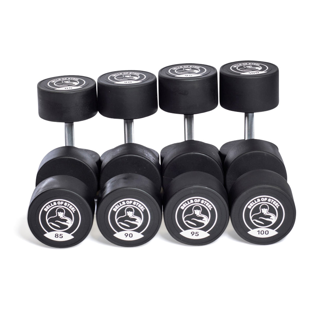 Five pairs of urethane dumbbells from Bells of Steel are arranged in a row, with weights labeled 85, 90, 95, and 100. Each boasts a logo on the end cap, highlighting their commercial-grade quality.
