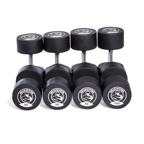 Five pairs of Bells of Steel Urethane Dumbbell Sets, labeled 85–100 lbs, are arranged in a row with circular logos on the ends—demonstrating commercial-grade quality for serious lifters.