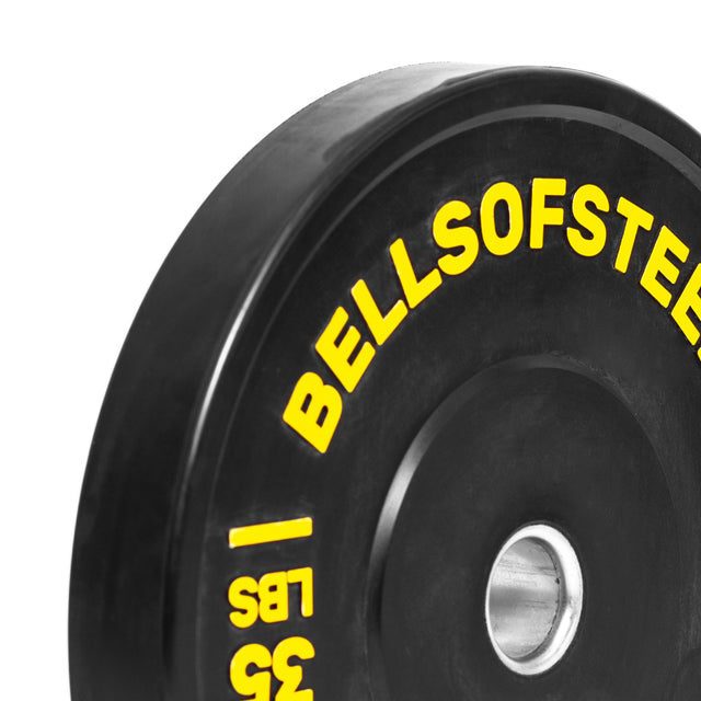 A close-up of a Bells of Steel Dead Bounce Conflict Bumper Plate Set in black, featuring yellow "BELLSOFSTEEL" and "35 LBS" text and a durable stainless steel insert ring at the center.