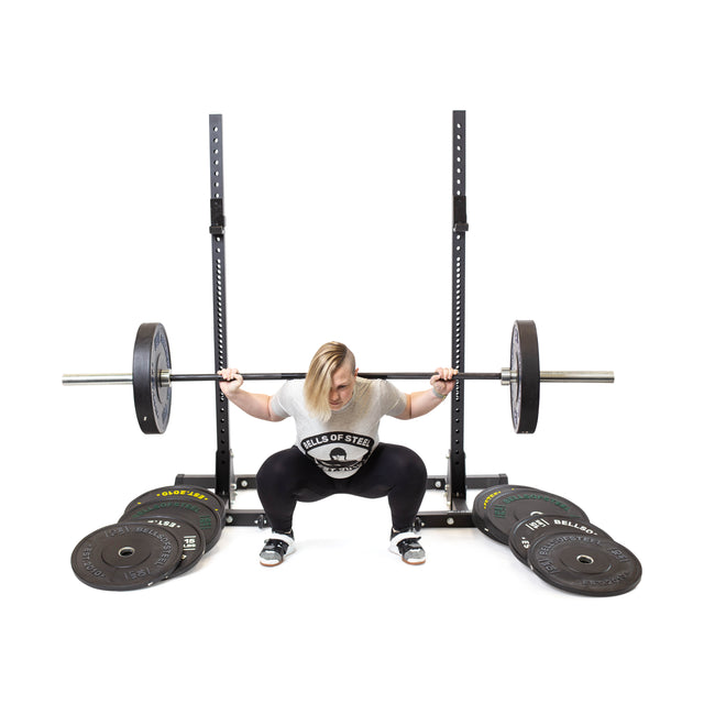 A blond-haired person in a Bells of Steel shirt performs a barbell squat in a squat rack. Dead Bounce Conflict Bumper Plate Sets by Bells of Steel are stacked on the floor nearby, all set against a completely white background.