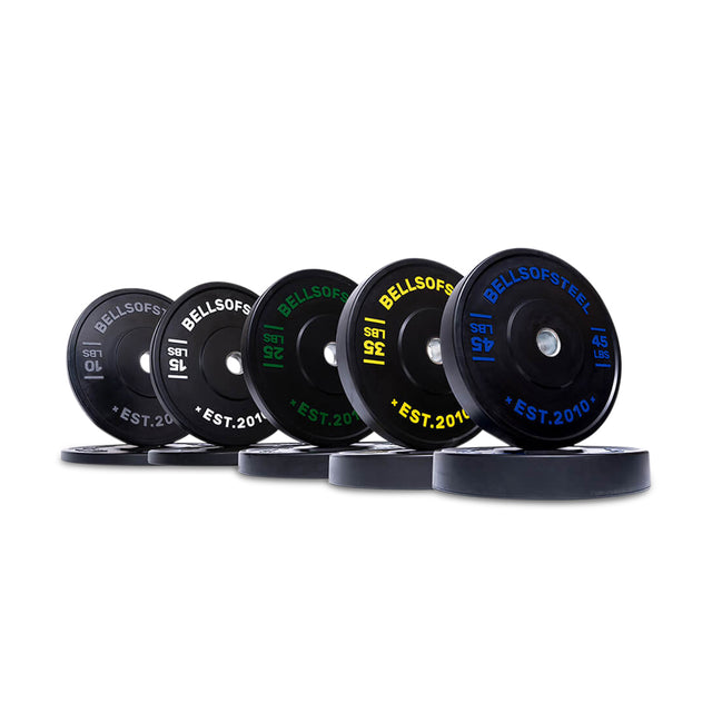Six color-coded Dead Bounce Conflict Bumper Plate Sets by Bells of Steel, each with a stainless steel insert, are arranged at an angle. Their black surfaces feature white, green, yellow, and blue text labeling weight in kilograms and pounds.
