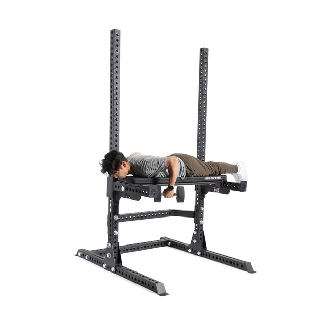 In a gym, someone uses a reverse hyperextension machine near a Bells of Steel Hydra/Manticore Utility Seat and a 3x3 rack, gripping the handles while lifting their legs behind them.