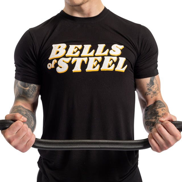 A person in a black Bells of Steel t-shirt grips the 54.5" EZ Curl Bar, showcasing tattooed forearms and arm gains, against a white background.