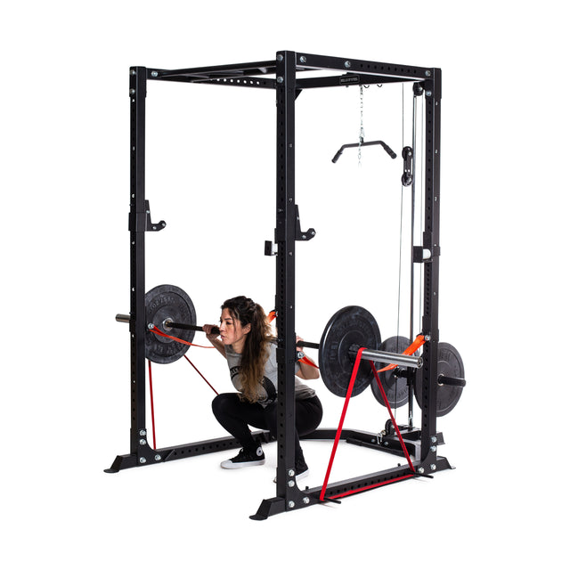 A woman squats inside the Bells of Steel Light Commercial Power Rack (2.3" x 2.3", ⅝" holes) with a barbell and resistance bands, making it ideal for safe home training.