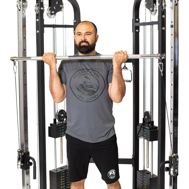 A bearded man in a gray t-shirt and black shorts stands inside a cable machine, gripping the Bells of Steel Double Hook Straight Bar Cable Attachment at chest height as if performing barbell exercises.