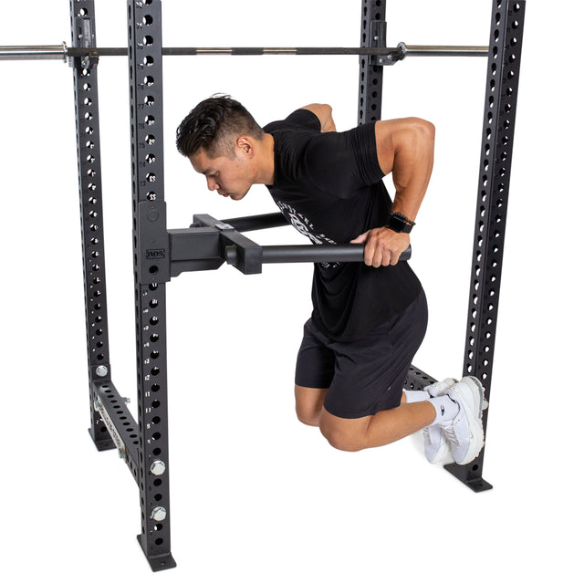 A man performs a dip exercise using a heavy-duty Bells of Steel Y Dip Bar Rack Attachment on parallel bars. He's wearing a black T-shirt, black shorts, and white sneakers, fully immersed in the focused gym atmosphere.