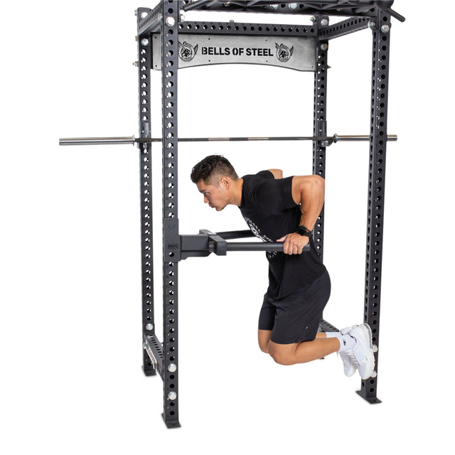 Dressed in black, a person does chest dips with bent knees using a Bells of Steel Y Dip Bar Rack Attachment for upper body strength. A barbell is set up overhead on the rack for squats.
