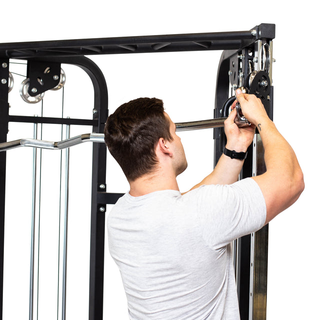 A man in a light gray t-shirt adjusts a Bells of Steel Double Hook EZ Curl Bar Cable Attachment on a cable machine, set against a clean, white background.