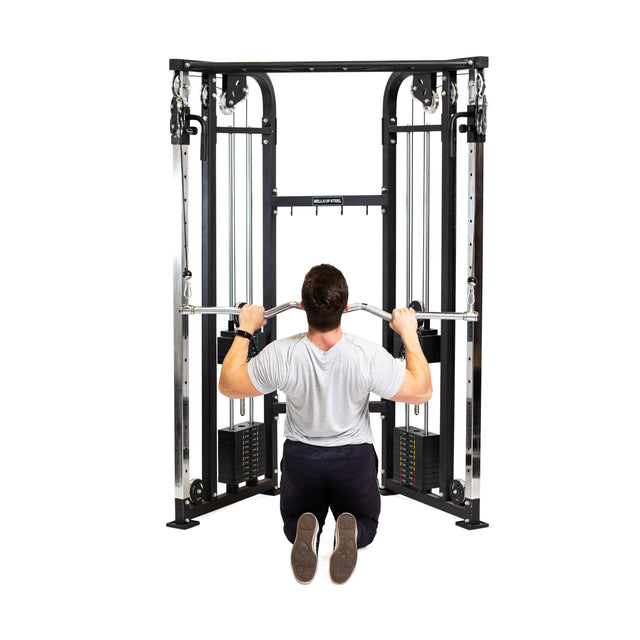 A person kneels on the floor facing a cable machine, gripping the Bells of Steel Double Hook EZ Curl Bar Cable Attachment with both hands, preparing to perform a joint-friendly back exercise.