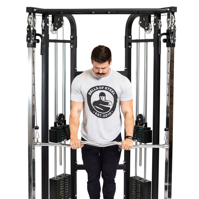 A man wearing a light gray "Bells of Steel" t-shirt and black pants uses the Bells of Steel Double Hook EZ Curl Bar Cable Attachment on a cable machine, standing against a white background.