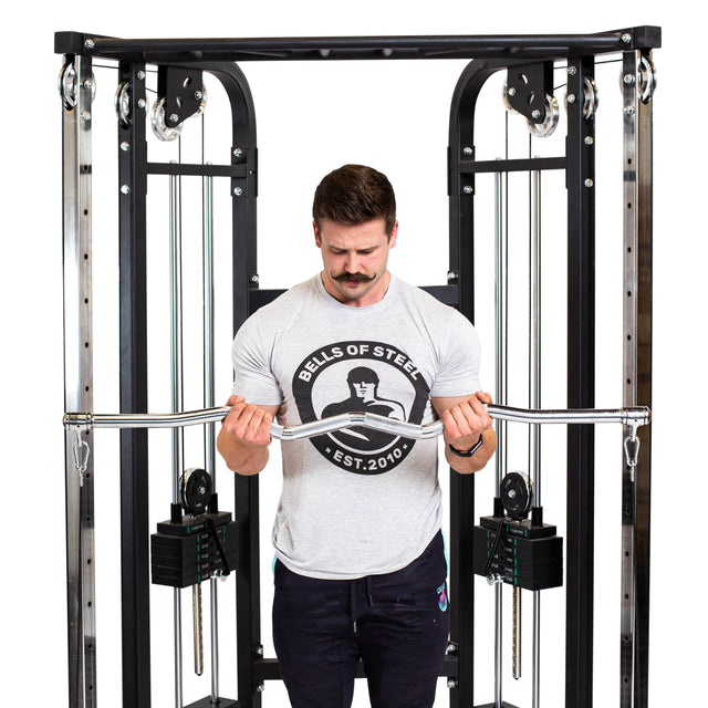 A man in a Bells of Steel t-shirt performs a joint-friendly barbell exercise using the Bells of Steel Double Hook EZ Curl Bar Cable Attachment on a cable machine with visible weight stacks.