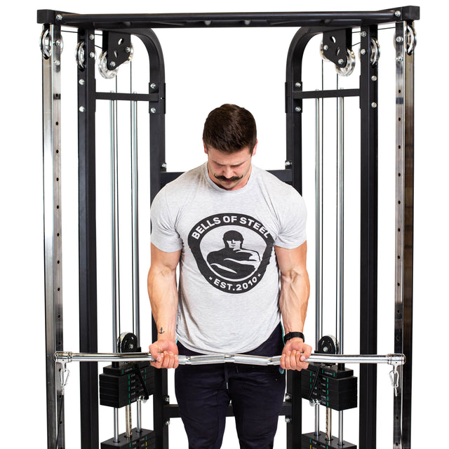 A man with a mustache performs a joint-friendly barbell exercise on a cable machine using the Bells of Steel Double Hook EZ Curl Bar Cable Attachment. He wears a light gray Bells of Steel T-shirt and black pants, focusing on his form.