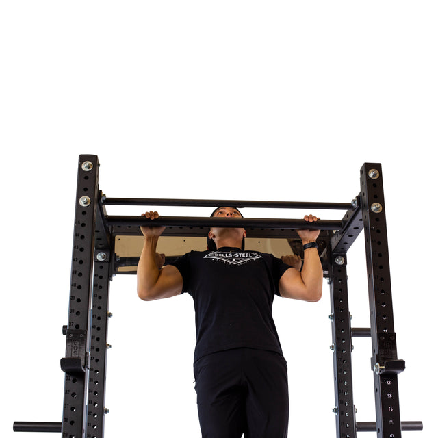 A person in a black t-shirt and pants performs a pull-up on the Bells of Steel Fat/Skinny Pull Up Bar, attached to a power rack, against a white background.