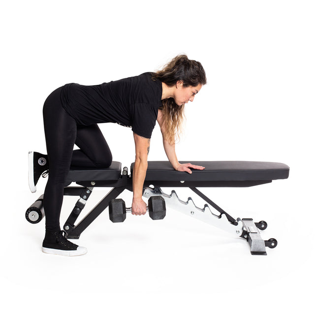 A woman in black workout clothes performs a one-arm row with a Bells of Steel Ergo Rubber Hex Dumbbell Set on a black adjustable bench, supporting herself with one knee and hand against a white background.