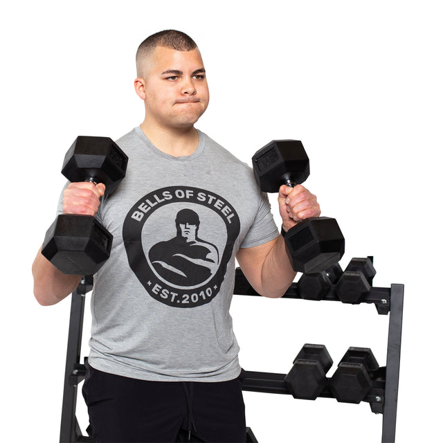 A man in a gray Bells of Steel T-shirt performs curls with Ergo Rubber Hex Dumbbell Sets by Bells of Steel, standing before a rack of dumbbells against a white background.