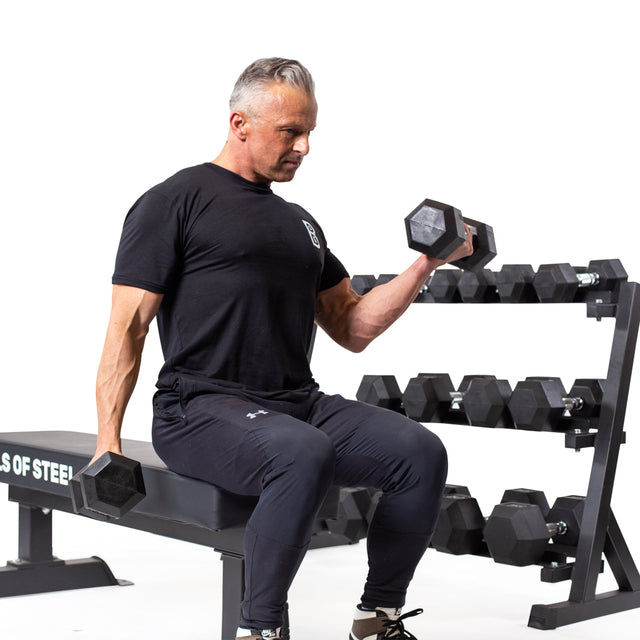 A man in a black t-shirt and pants sits on a bench, performing bicep curls with Bells of Steel Ergo Rubber Hex Dumbbell Sets. A rack of these hex dumbbells is visible in the background against a white backdrop.
