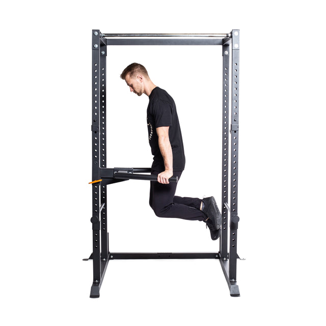 An individual is performing a tricep dip using the Bells of Steel Y Dip Bar Rack Attachment, gripping the bars on a metal power rack with legs bent and feet elevated.