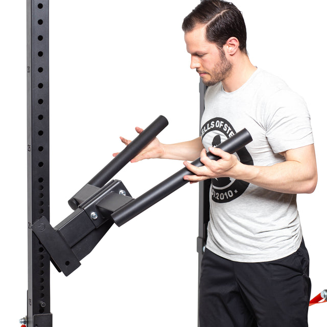 Clad in a gray t-shirt, a man adjusts the detachable arms of a Bells of Steel gym power rack with the Y Dip Bar Rack Attachment. He ensures the black sturdy equipment can support its 500lb capacity against a plain white background.