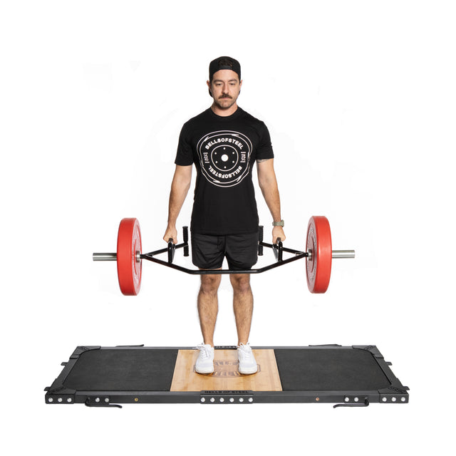 A man on a Bells of Steel Weightlifting Platform grips an Olympic barbell with red weights. He's wearing a black cap, black t-shirt with a circular logo, black shorts, and white shoes, gazing downward with a firm stance.