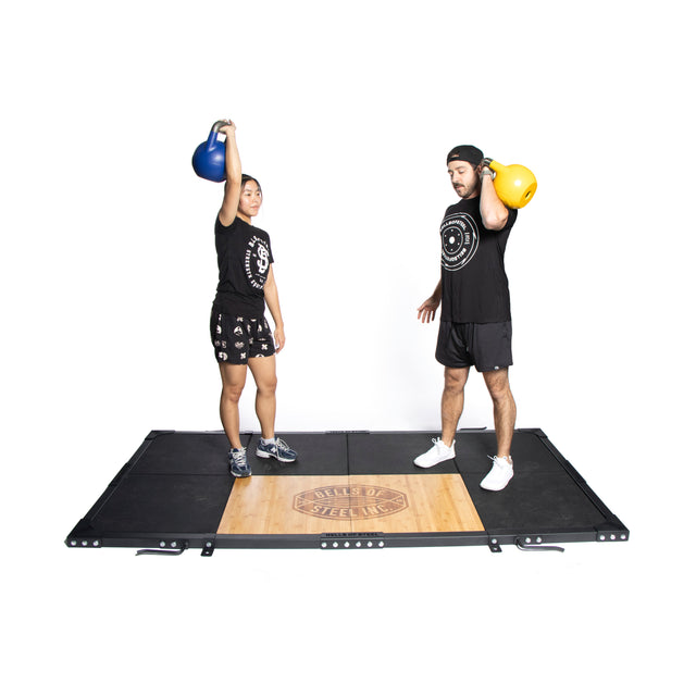 Two people stand on a Bells of Steel Weightlifting Platform, both in black athletic wear. The left person lifts a blue kettlebell overhead while the right holds a yellow one at shoulder height, surrounded by deadlift pads and an Olympic barbell.